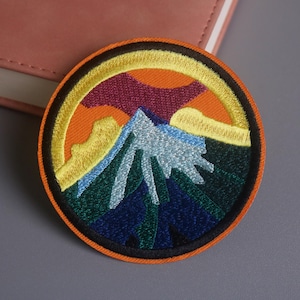 May include: Embroidered patch featuring a mountain scene with a blue peak, orange sky, and yellow accents. The patch is circular with a black border. The design evokes a sense of nature and adventure.