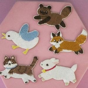 May include: Five embroidered iron-on patches featuring various animals: a brown raccoon, a white bird, a brown and white cat, a white lamb, and an orange fox.