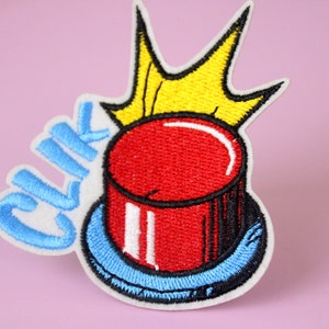 May include: A red button with a white rim and a blue base, with the word "Click" embroidered in blue on the side. A yellow crown is above the button.