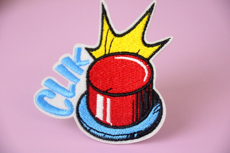 May include: A red button with a white rim and a blue base, with the word "Click" embroidered in blue on the side. A yellow crown is above the button.