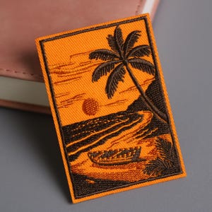 Op de afbeelding: Geborduurde patch met een zonsondergangscène met een palmboom, oceaan en een boot. De patch heeft een oranje achtergrond met bruine details. De patch is rechthoekig en heeft een zwarte rand.