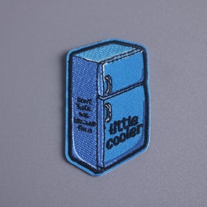 May include: A blue embroidered patch shaped like a fridge, with the words "little cooler" and "don't safe me because I'm fresh" in black. The patch is edged in a darker blue, creating a defined border.