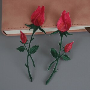以下が含まれることがあります： 緑色の茎と葉が付いた赤い刺繍のバラのパッチが2つ。パッチは灰色の表面にあります。