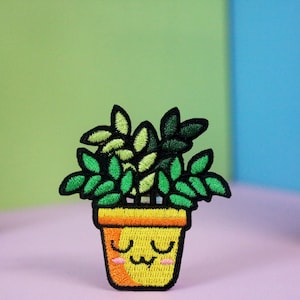 Puede incluir: Un parche bordado que representa una planta en maceta con una cara sonriente. La planta tiene varias hojas y está en una maceta amarilla con un borde naranja.