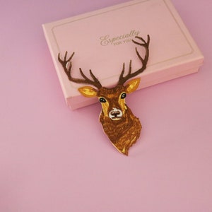 May include: A brown deer head decoration with antlers, placed on a pink gift box. The deer head is detailed with yellow and brown tones. The gift box is light pink with the words "Especially FOR YOU" in gold.
