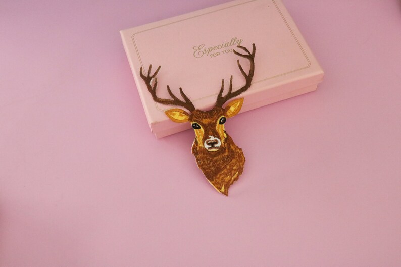 May include: A brown deer head decoration with antlers, placed on a pink gift box. The deer head is detailed with yellow and brown tones. The gift box is light pink with the words "Especially FOR YOU" in gold.