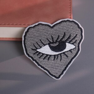 May include: A gray heart-shaped embroidered patch featuring a stylized eye with long eyelashes. The eye is black and white, and the patch has a white border. The patch is placed on a gray surface.