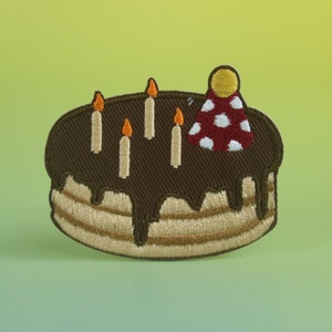 Peut inclure: Un patch brodé représentant un gâteau au chocolat avec un glaçage blanc et cinq bougies allumées. Le gâteau est surmonté d'un chapeau de fête à pois rouges et blancs.