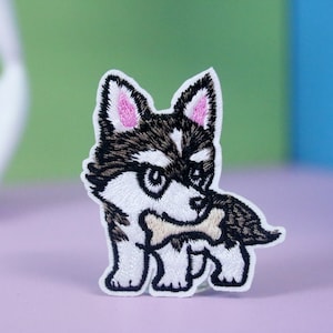 May include: Embroidered iron-on patch of a cartoon husky puppy with black and white fur, pink ears, and holding a bone in its mouth.