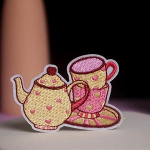 Könnte beinhalten: Gestickter Aufnäher mit einem Teekannen- und Tassenset. Die Teekanne ist gelb mit roten Herzakzenten und einem roten Deckel. Die Tasse ist rosa mit gelben Herzakzenten. Der Aufnäher ist weiß umrandet.
