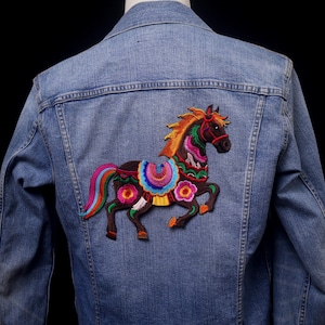May include: A light blue denim jacket with a colourful embroidered horse design on the back. The horse features a brown body, a rainbow mane and tail, and floral accents in pink, orange, and blue.