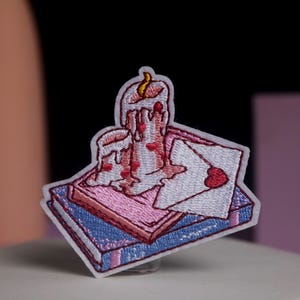 Può includere: Toppa ricamata raffigurante una candela con cera che si scioglie e un disegno a forma di cuore, sopra una pila di libri e una busta. La toppa ha un bordo bianco, con accenti rosa, rossi e blu. Il design è dettagliato e colorato.