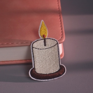 May include: An embroidered patch shaped like a lit candle. The candle is light gray with a dark brown base and a yellow flame with a small red tip. The patch has a white border and is positioned against a pink rectangular object with rounded corners.