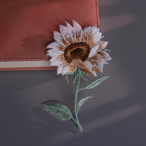 Puede incluir: Parche de girasol bordado con pétalos blancos, centro marrón y tallo y hojas verdes. El parche se coloca sobre una superficie gris junto a un cuaderno de cuero rosa.