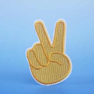 Puede incluir: Un parche bordado de color amarillo en forma de signo de paz con costuras blancas.