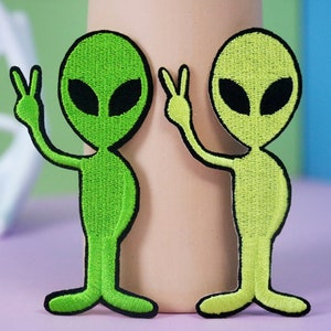 Puede incluir: Dos parches bordados de extraterrestres verdes con ojos negros y un signo de paz. El extraterrestre de la izquierda es de un verde más oscuro y el extraterrestre de la derecha es de un verde más claro.