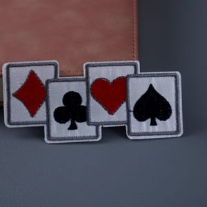 Könnte beinhalten: Vier Bügelbilder mit den vier Kartenfarben: eine rote Raute, ein schwarzer Klee, ein rotes Herz und ein schwarzer Pik. Die Patches sind weiß mit einem grauen Rand.