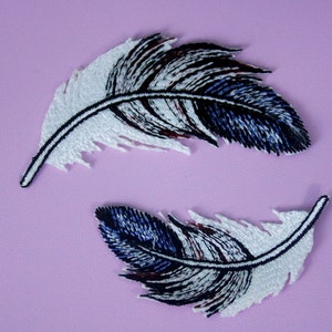 Puede incluir: Dos parches de plumas bordadas en blanco y azul. Las plumas están dispuestas en círculo, con las puntas de las plumas apuntando hacia el centro del círculo.