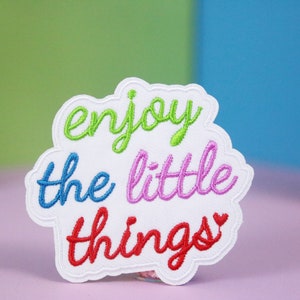 May include: White embroidered patch with the phrase "enjoy the little things" in rainbow colors. A small red heart is at the end of the phrase.