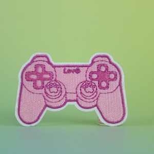 May include: Pink embroidered patch of a video game controller. The controller has a white outline and pink buttons.