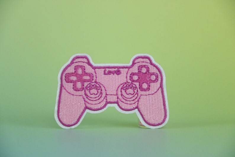 May include: Pink embroidered patch of a video game controller. The controller has a white outline and pink buttons.