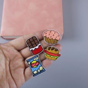 May include: Four embroidered iron-on patches featuring a chocolate bar, a cupcake, a hamburger, and a bag of chips.