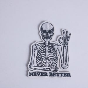 Puede incluir: Parche bordado en blanco y negro con un esqueleto haciendo el gesto de la mano de 'ok'. El parche tiene las palabras 'NEVER BETTER' en texto negro en la parte inferior. El parche está sobre un fondo blanco.
