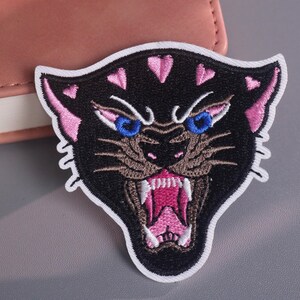 Könnte beinhalten: Gestickter Aufnäher mit einem schwarzen Pantherkopf mit rosa Akzenten und blauen Augen. Der Panther öffnet sein Maul und zeigt rosa Zähne und Zunge. Der Aufnäher hat einen weißen Rand.