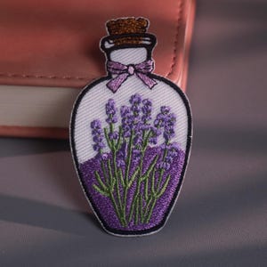 May include: Embroidered patch featuring a lavender potion bottle. The patch has a white background with a purple bottle shape, a cork top, and a purple bow. Inside the bottle are purple lavender flowers with green stems.