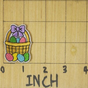 Peut inclure: &Eacute;cusson brod&eacute; d'un panier de P&acirc;ques avec un n&oelig;ud violet, rempli d'&oelig;ufs color&eacute;s. Le patch mesure environ 2,5 cm de hauteur et de largeur. Le patch est sur une surface en bois avec des marquages en pouces.