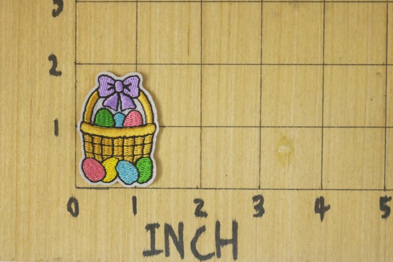 Peut inclure: &Eacute;cusson brod&eacute; d'un panier de P&acirc;ques avec un n&oelig;ud violet, rempli d'&oelig;ufs color&eacute;s. Le patch mesure environ 2,5 cm de hauteur et de largeur. Le patch est sur une surface en bois avec des marquages en pouces.