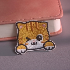 May include: An embroidered patch featuring a winking orange tabby cat with dark orange stripes, a white muzzle, and two paws resting at the bottom. The cat has pink inner ears, a small pink nose, and a black outline, creating a detailed feline design.