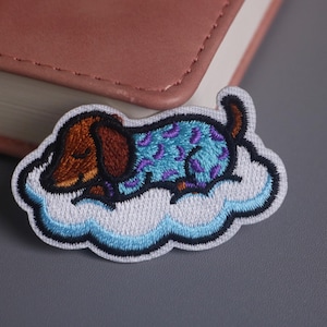 May include: Embroidered patch featuring a sleeping dachshund dog on a cloud. The dog has brown and tan fur with a blue and purple patterned outfit. The cloud is white with a blue outline. The patch is designed for sewing or ironing onto fabric.