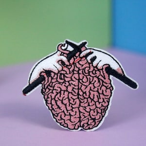 Könnte beinhalten: Ein gestickter Bügel-Patch mit einem rosa Gehirn, das von zwei Händen mit Nadeln zusammengenäht wird. Das Gehirn ist mit einer realistischen Textur detailliert.