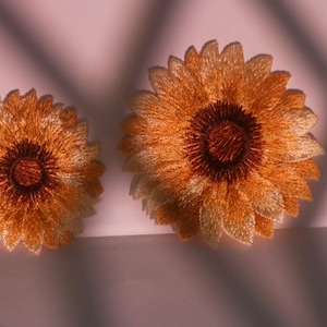 May include: Two embroidered sunflower patches. The sunflowers have a detailed design with yellow and orange petals and a dark brown centre. The patches are on a light pink surface with shadows.