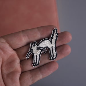 Può includere: Toppa ricamata raffigurante un gatto stilizzato in bianco con contorno nero. Il gatto è arcuato con la coda sollevata. La toppa è tenuta in mano su sfondo neutro. Per abbigliamento o accessori.