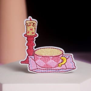 May include: Embroidered patch showing a pink teacup with a yellow handle on a pink and blue embroidered napkin. A red candle and candlestick are to the left. The candle has a yellow flame and red heart details.