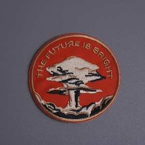 Op de afbeelding: Geborduurde patch met een rode achtergrond en de tekst "THE FUTURE IS BRIGHT" in een cirkelvormig ontwerp. De patch heeft een gestileerde paddenstoelwolk-afbeelding in wit, zwart en bruin.