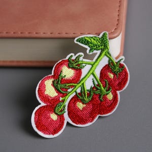 May include: Embroidered patch featuring a cluster of red tomatoes with green stems and leaves. The tomatoes have a textured appearance with yellow highlights. The patch is outlined in white.
