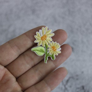 Peut inclure: Un patch brodé de fleurs jaunes et blanches avec des feuilles vertes.