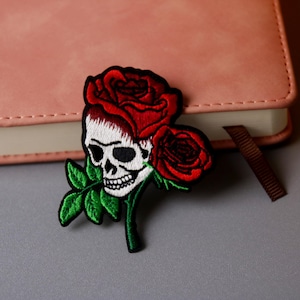 Puede incluir: Parche bordado con una calavera blanca adornada con una corona de rosas rojas y dos rosas rojas adicionales. Hojas verdes y un borde negro completan el diseño. El parche está sobre un cuaderno rosa.