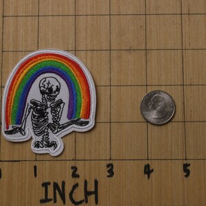 以下が含まれることがあります： 虹の下に座っている骸骨が描かれた白い刺繍パッチ。虹は赤、オレンジ、黄、緑、青、藍、紫の糸でできています。パッチの幅は約5cmです。