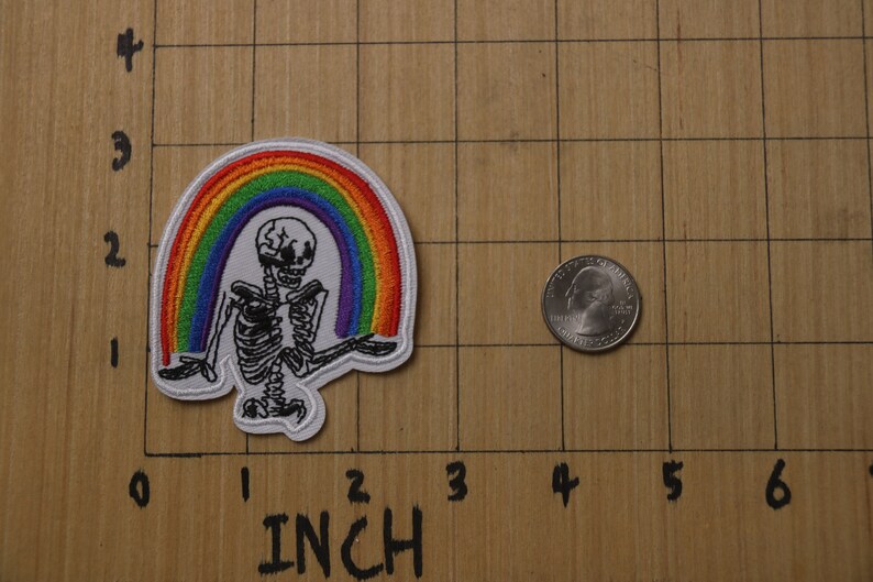 以下が含まれることがあります： 虹の下に座っている骸骨が描かれた白い刺繍パッチ。虹は赤、オレンジ、黄、緑、青、藍、紫の糸でできています。パッチの幅は約5cmです。