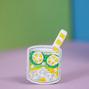 以下が含まれることがあります： 黄色と白のストライプのストローが付いたレモネードグラスの白い刺繍パッチ。レモネードは緑と黄色で、白い氷とレモンのスライスが2つ入っています。