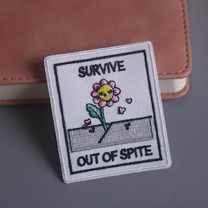 May include: Embroidered patch with the words "SURVIVE OUT OF SPITE" above and below a cartoon flower with a pink face and yellow center. The patch is white with a black border and is rectangular.