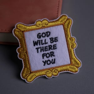 May include: Embroidered patch in the shape of a gold picture frame with the words "GOD WILL BE THERE FOR YOU" in black. The patch is on a white background and has a decorative border.