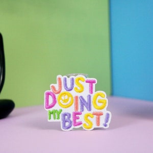 May include: A white embroidered patch with the text "Just Doing My Best!" in colorful letters. A yellow smiley face is included in the word "Doing".