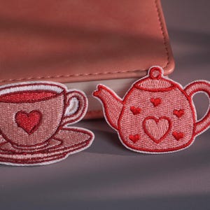 May include: Embroidered patches of a teacup and saucer, and a teapot, in pink and red hues. Each patch features a heart design. The teacup has a red heart in the centre. The teapot has a large heart and smaller hearts embroidered on it.