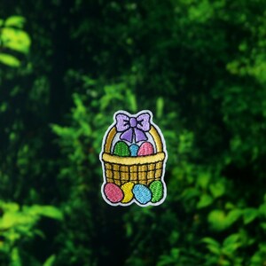 Peut inclure: &Eacute;cusson brod&eacute; repr&eacute;sentant un panier jaune rempli d'&oelig;ufs de P&acirc;ques color&eacute;s. Le panier est orn&eacute; d'un n&oelig;ud violet et bord&eacute; de blanc. Les &oelig;ufs sont roses, bleus, verts et violets. L'&eacute;cusson est pos&eacute; sur un fond vert.