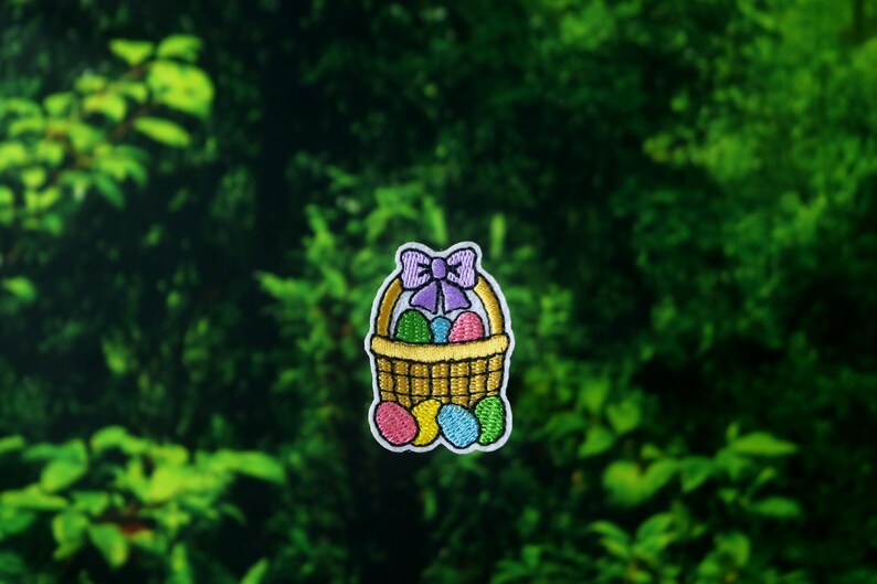 Peut inclure: &Eacute;cusson brod&eacute; repr&eacute;sentant un panier jaune rempli d'&oelig;ufs de P&acirc;ques color&eacute;s. Le panier est orn&eacute; d'un n&oelig;ud violet et bord&eacute; de blanc. Les &oelig;ufs sont roses, bleus, verts et violets. L'&eacute;cusson est pos&eacute; sur un fond vert.